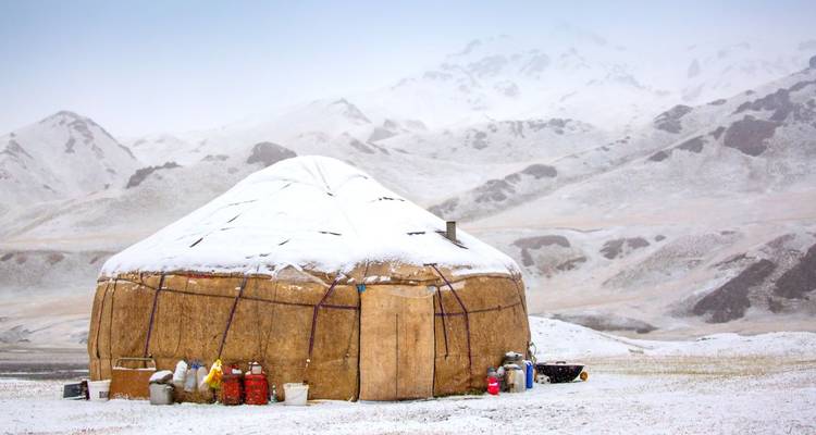 A traditional yurt covered in snow against a mountainous backdrop.
