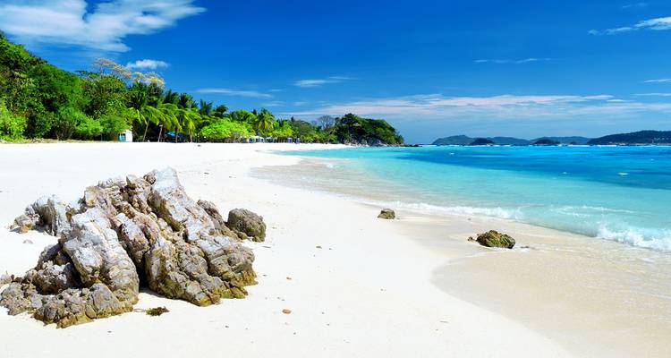 Unberührter tropischer Strand mit weißem Sand, eine Felsenformation im Vordergrund, türkisfarbenes Meer und tiefblauer Himmel