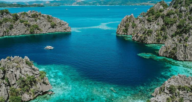Luftaufnahme einer leuchtend blauen Lagune, umgeben von zerklüfteten Kalksteinfelsen mit einem einsamen Banca-Boot in Coron