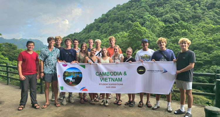 Gran grupo de expedición estudiantil posando con una pancarta de Camboya y Vietnam contra un exuberante telón de fondo montañoso verde.