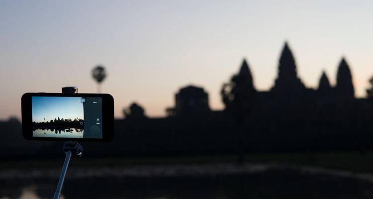 Teléfono inteligente en un palo de selfie capturando la silueta del amanecer de Angkor Wat a través de un estanque reflectante.