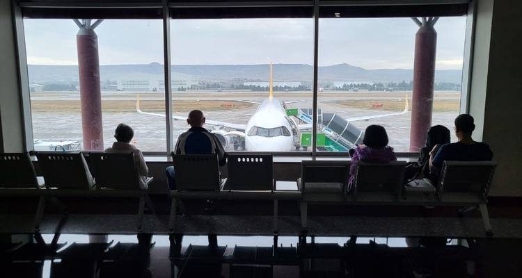 Mensen zittend in een wachtruimte op een luchthaven, vliegtuig zichtbaar buiten.