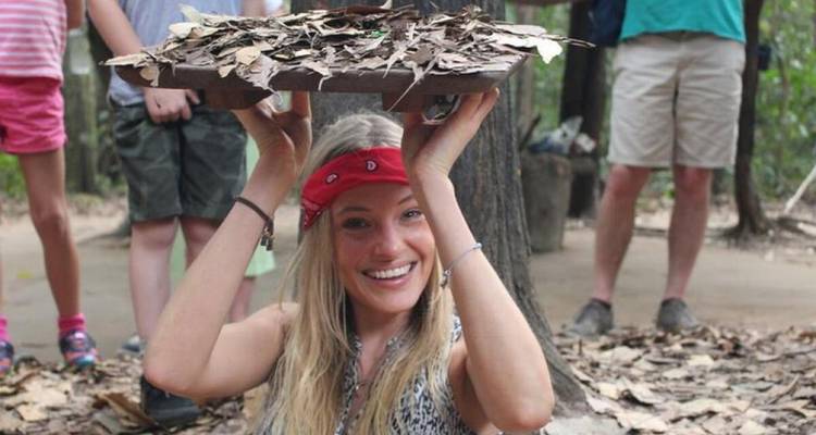 Smiling person holding up a leaf-covered lid at a tunnel entrance.