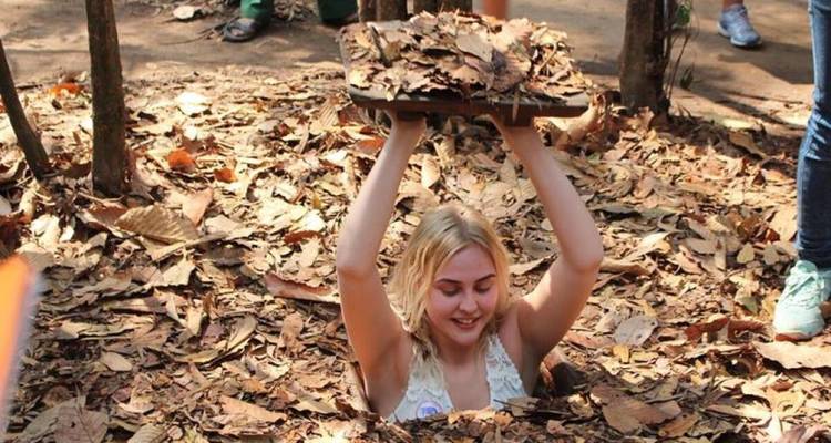 Smiling woman holding a leaf-covered lid while climbing out of a tunnel.