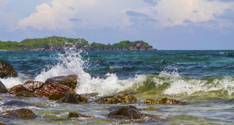 Des vagues qui se brisent contre les rochers avec une vue lointaine d'une île verdoyante et luxuriante.