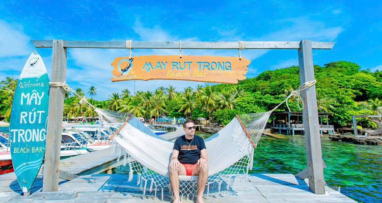 Een man ontspant zich in een witte hangmat op een houten steiger naast een turquoise zee onder een rustiek strandbar-bord op Phu Quoc.