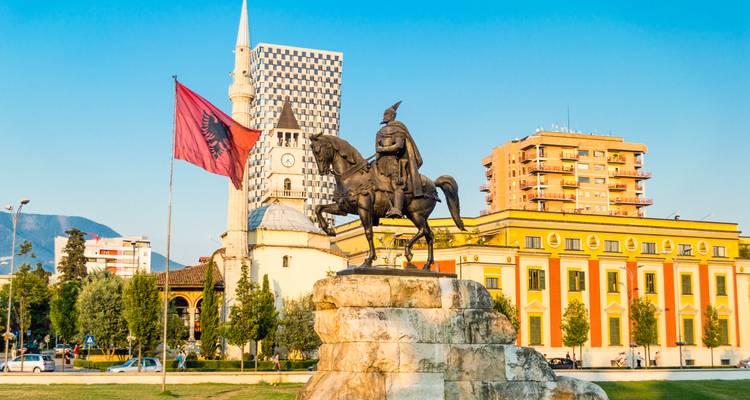 Ruiterstandbeeld van Skanderbeg, Albanese vlag en kleurrijke regeringsgebouwen gloeien onder warm avondlicht op het hoofdplein van Tirana.