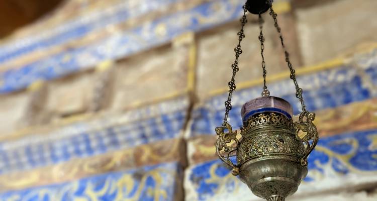 Close-up van een sierlijke hangende metalen lamp in een ingewikkeld gedecoreerd historisch gebouw met een wazige tegelwerkachtergrond.