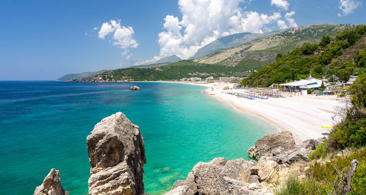 Turquoise strand met wit zand, rotsachtige voorgrond en groene bergen onder een heldere blauwe hemel