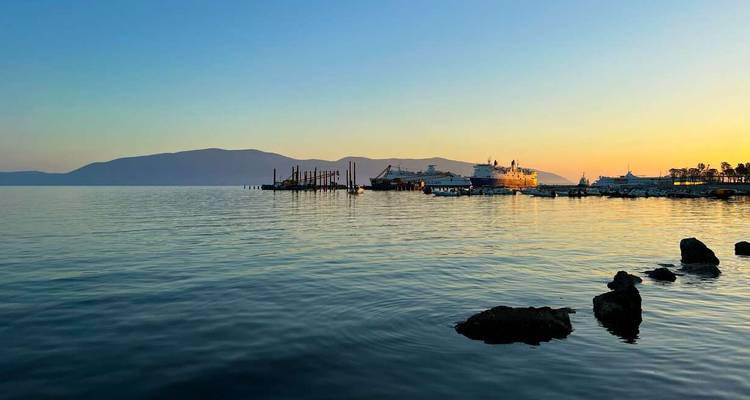 Kalme zee bij schemering met veerboten afgemeerd naast een pier en zacht oranje licht op verre heuvels.
