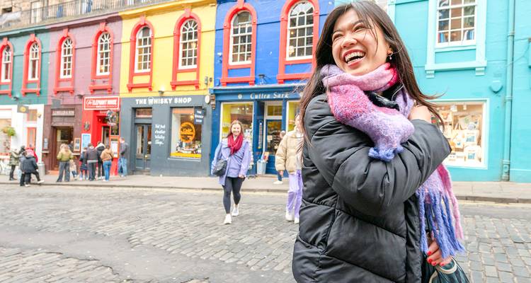 Mujer joven sonriente en una calle empedrada con escaparates de tiendas pintados de colores brillantes y transeúntes.