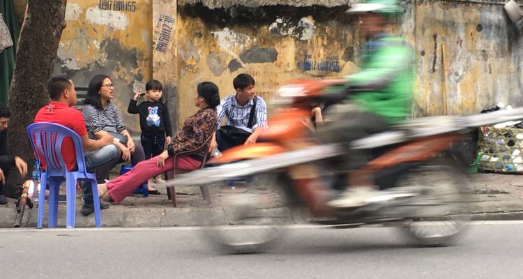 Les habitants discutent sur des tabourets en plastique près d'un mur vieilli tandis qu'une moto floue passe à toute vitesse.