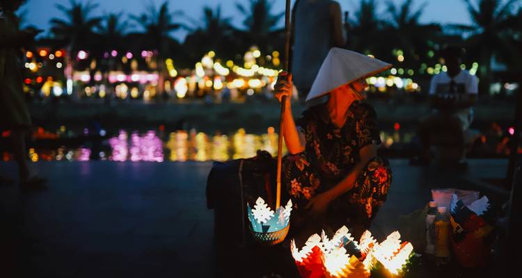 Vendeur de lanternes au chapeau conique s'agenouille à côté de lanternes en papier lumineuses sur un bord de rivière la nuit.