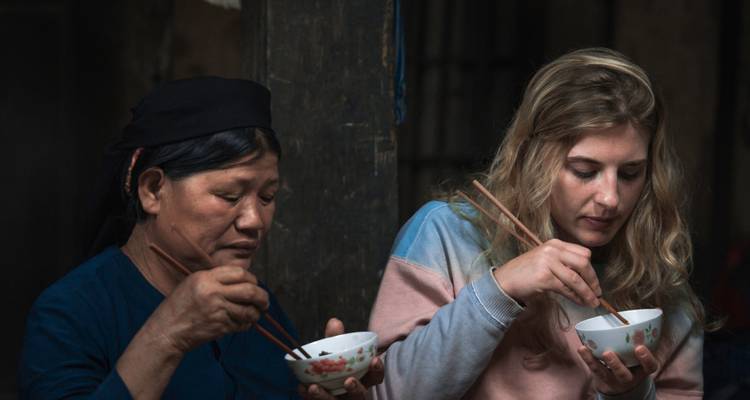 Une femme locale et un touriste partagent un repas simple avec des baguettes à l'intérieur d'une maison rustique.