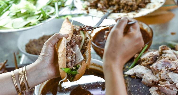 Un vendeur prépare une baguette banh mi au-dessus de bols de viande et d'ingrédients pour la sauce.