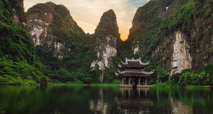 Une pagode délicate se dresse sur pilotis dans un lac tranquille entouré de falaises calcaires escarpées au coucher du soleil.