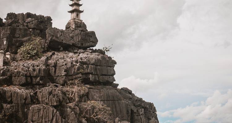 Une pagode de pierre se dresse au sommet de rochers karstiques gris et escarpés sous un ciel nuageux à Ninh Binh.