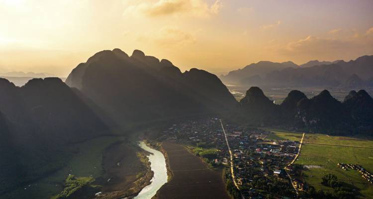 Une vue aérienne panoramique d'une vallée rurale vietnamienne où une rivière serpente près d'un petit village sous des montagnes karstiques spectaculaires baignées dans la lumière dorée et chaleureuse de l'heure dorée.