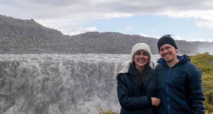 Glimlachend stel in winterjassen poseert naast een krachtige grijze waterval omringd door rotsige kliffen