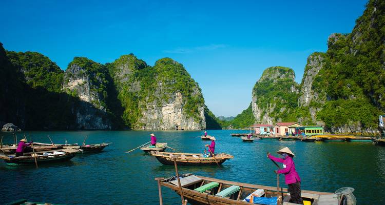 Des femmes en violet rament dans des sampans en bois à travers les eaux émeraude d'une baie calcaire sous un ciel dégagé.