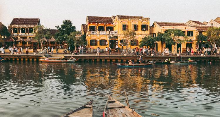Scène au bord de l'eau à Hoi An avec des bâtiments patrimoniaux jaunes, des bateaux et des foules reflétés dans une eau calme au crépuscule.