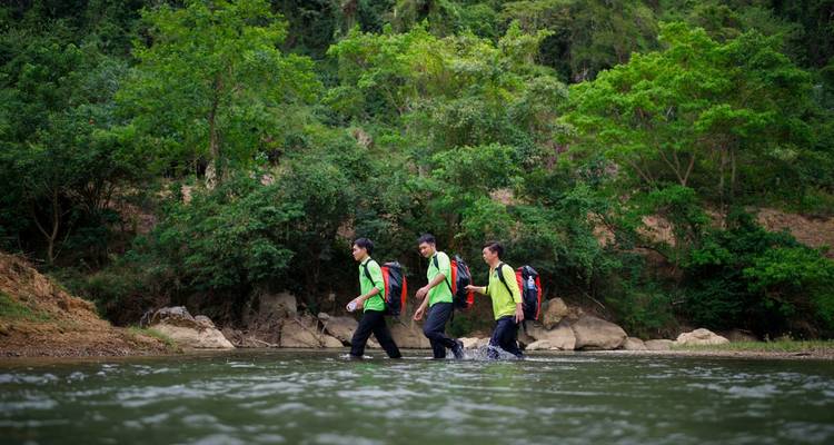 Trois randonneurs traversent à gué une rivière de jungle peu profonde en portant des gilets de sécurité colorés et des sacs à dos.