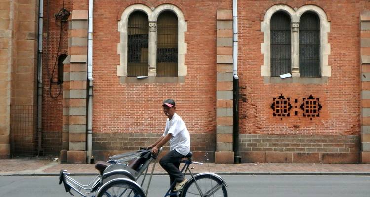 Un homme pédale sur un cyclo-pousse traditionnel devant un bâtiment colonial en briques rouges avec des fenêtres cintrées.