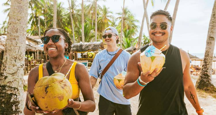 Viajeros sonrientes caminan por una playa bordeada de cocoteros sosteniendo cocos recién cortados.