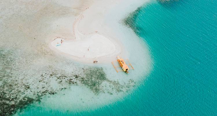 Vista aérea de un pequeño islote de arena blanca con una embarcación con balancín en agua turquesa cristalina.