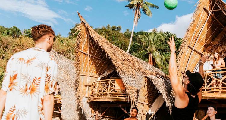 Des voyageurs jouent avec un ballon devant des huttes de plage en bambou sous les palmiers.