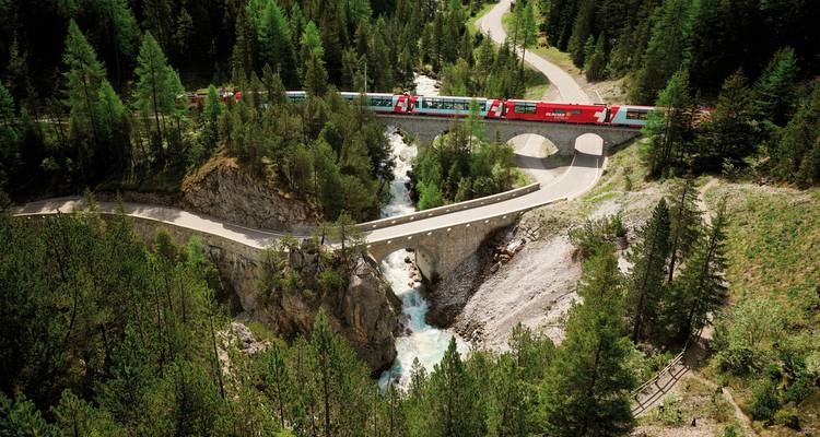Scenic view of a red train crossing a bridge over a river gorge surrounded by lush greenery.