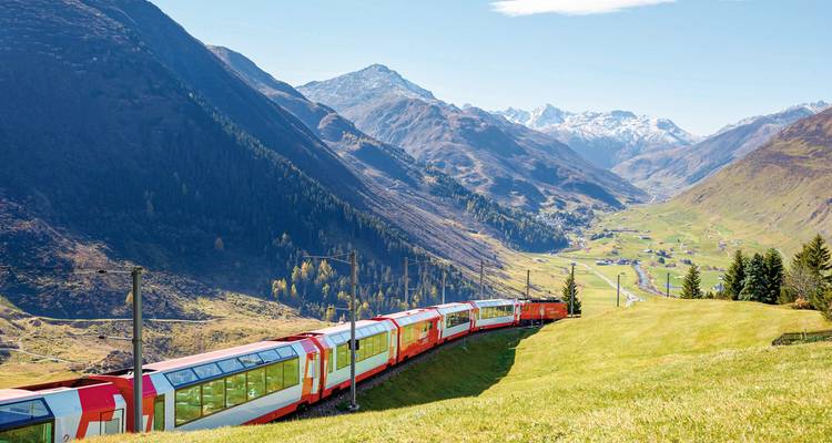 A red train traveling through a picturesque mountain valley with clear skies.