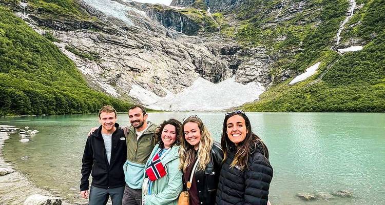 Grupo de excursionistas sonriendo junto a un lago glacial turquesa rodeado de empinadas montañas noruegas y parches de nieve.