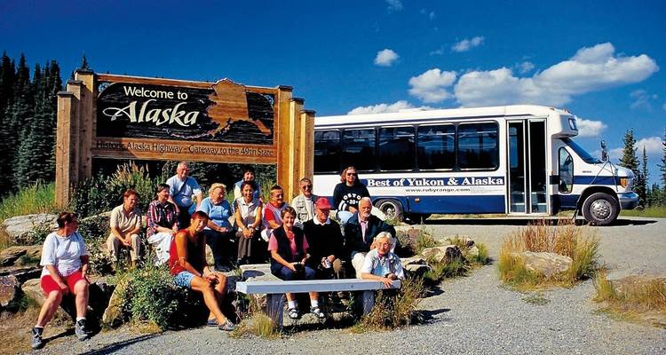 Tour group poses by a ‘Welcome to Alaska’ sign beside their tour bus on a sunny day.