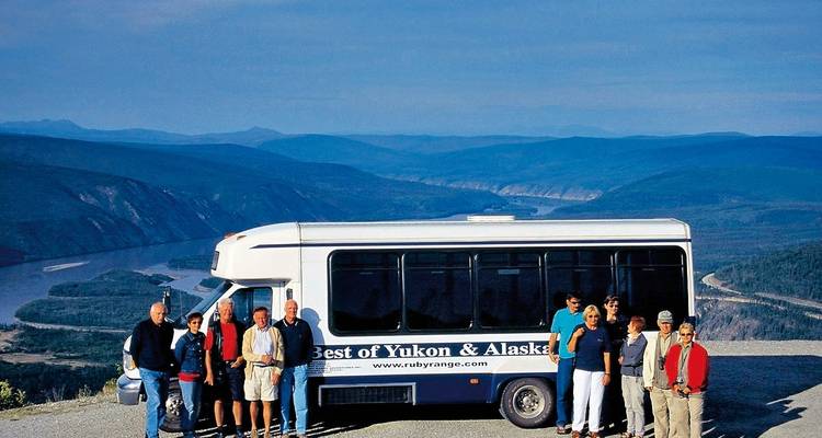 Travelers stand beside a tour bus overlooking a vast winding river valley in Alaska or Yukon.