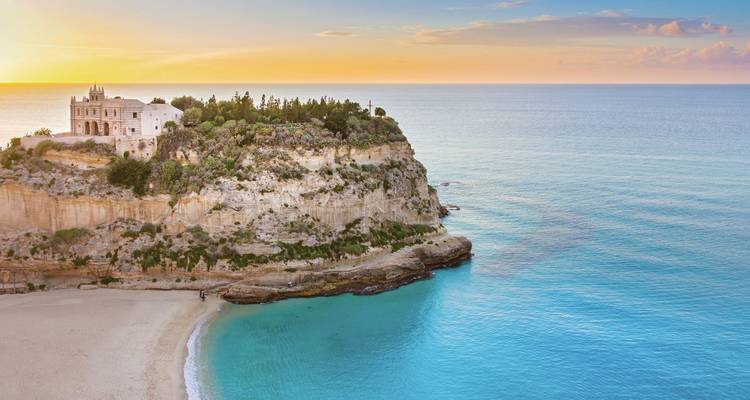 Sanctuaire au sommet de la falaise et plage à Tropea éclairés par un coucher de soleil coloré sur une mer turquoise.