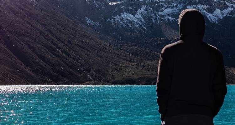 Een persoon in een donkere hoodie die over een turquoise meer naar bergen kijkt.