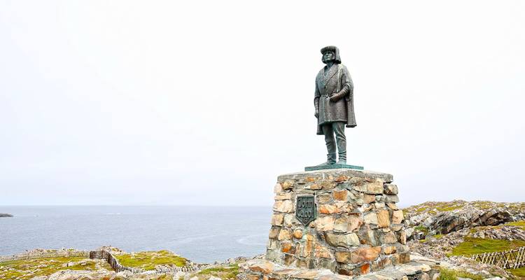 Statue de bronze d'un explorateur sur un socle de pierre dominant un cap côtier accidenté et une mer grise.