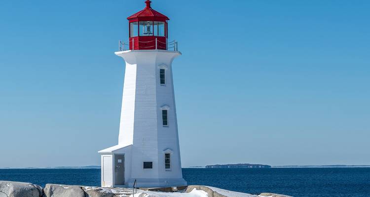 A white lighthouse with a red top on a rocky shore under a clear blue sky.