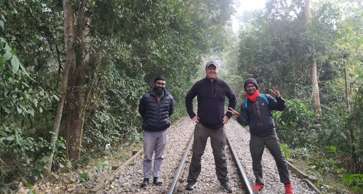 Tres amigos están parados en las vías del tren que atraviesan un bosque verde y denso, sonriendo a la cámara.