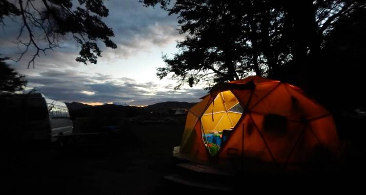 Leuchtend gelbes geodaetisches Zelt auf einem dunklen Campingplatz mit silhouettierten Baeumen und Dämmerungshimmel