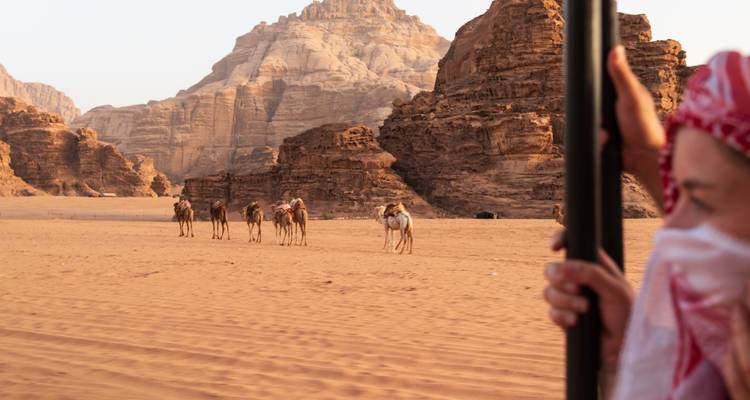 Camel caravan traverses vast sandy expanse backed by towering rock mountains in Wadi Rum.