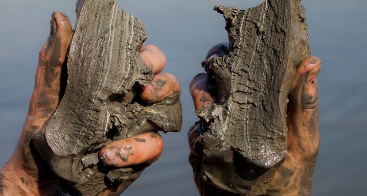 Close-up of muddy hands holding thick dark mineral mud over calm water