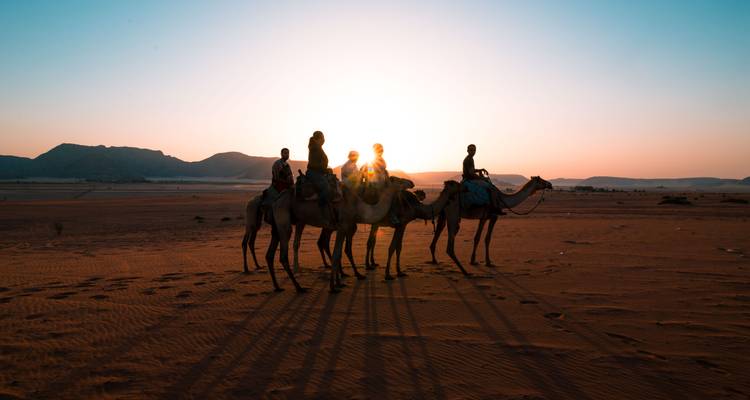 Silhouettes of travelers riding camels across the red desert at sunrise with long shadows.