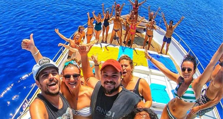 A lively group of young travelers posing on the bow of a boat in vibrant blue water, arms raised in excitement.