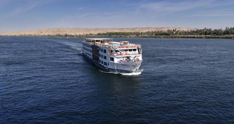 Luchtfoto van een groot wit Nijl-cruiseschip dat door diepblauw water glijdt met woestijnoevers in de verte.