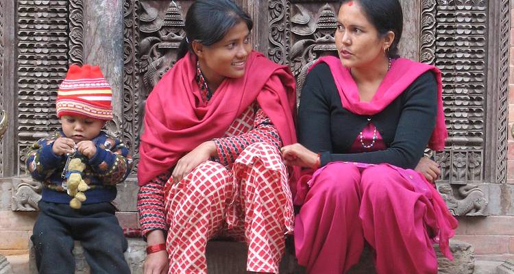 Frauen und Kind in traditioneller Kleidung sitzen neben kunstvollen Holzschnitzereien.