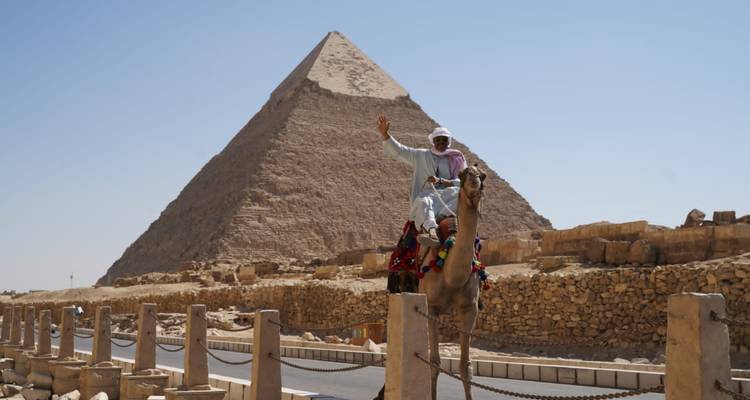 Persoon op kameel die zwaait met piramide in zicht.