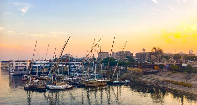 Uitzicht op een rivier met aangemeerde boten en gebouwen op de achtergrond tijdens zonsondergang.