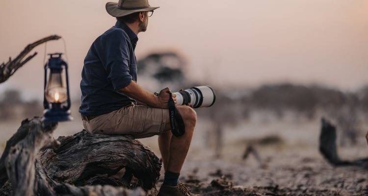 Mann sitzt auf einem Baumstamm mit einer Kamera in natürlicher Umgebung.